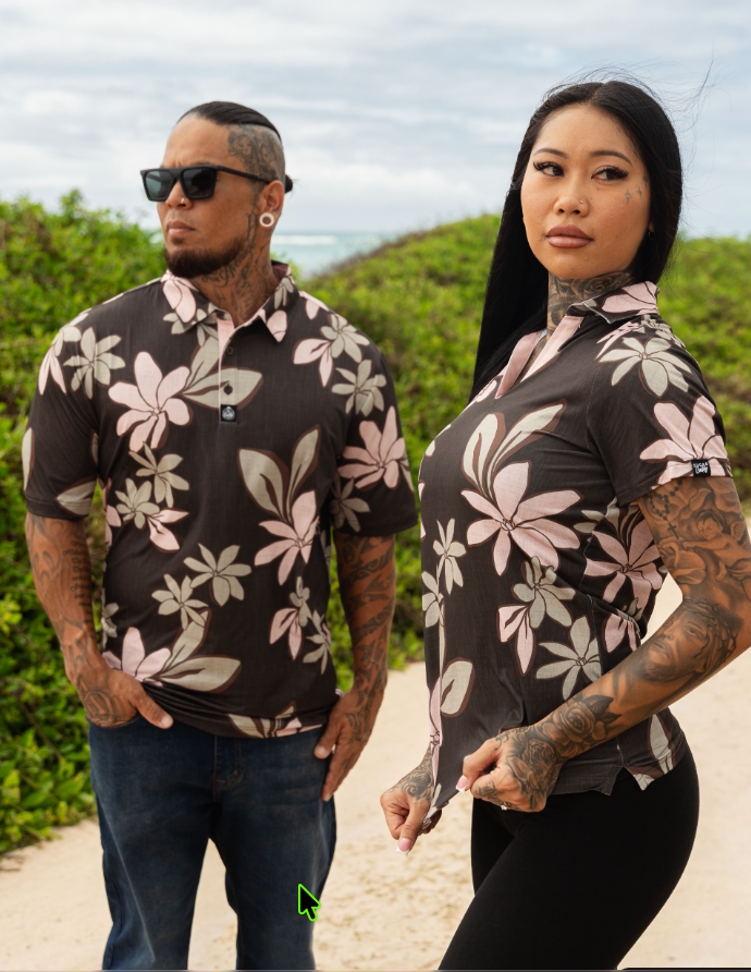 A man and a woman modeling matching short-sleeve button-up shirts with a dark brown base and a pink and beige floral pattern, set against a beach and greenery background.
