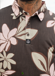Close-up of a men's short-sleeve polo shirt featuring a brown base with a large floral print in beige and light pink. The shirt has a three-button placket and a small black brand patch on the chest.