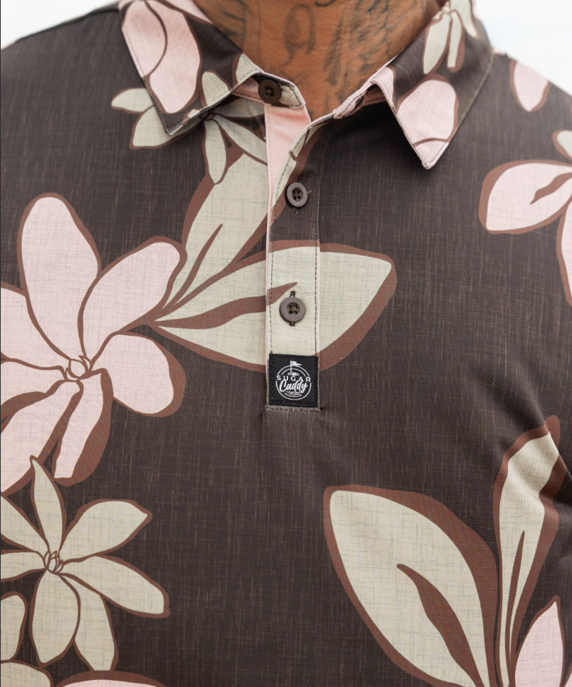 Close-up of a men's short-sleeve polo shirt featuring a brown base with a large floral print in beige and light pink. The shirt has a three-button placket and a small black brand patch on the chest.