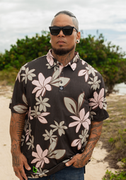 A man wearing a dark brown short-sleeve button-up shirt with a pink and beige floral print, styled with sunglasses and a beard, standing outdoors on a sandy path with green foliage in the background.