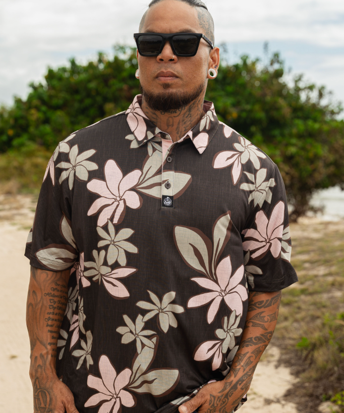 A man wearing a dark brown short-sleeve button-up shirt with a pink and beige floral print, styled with sunglasses and a beard, standing outdoors on a sandy path with green foliage in the background.