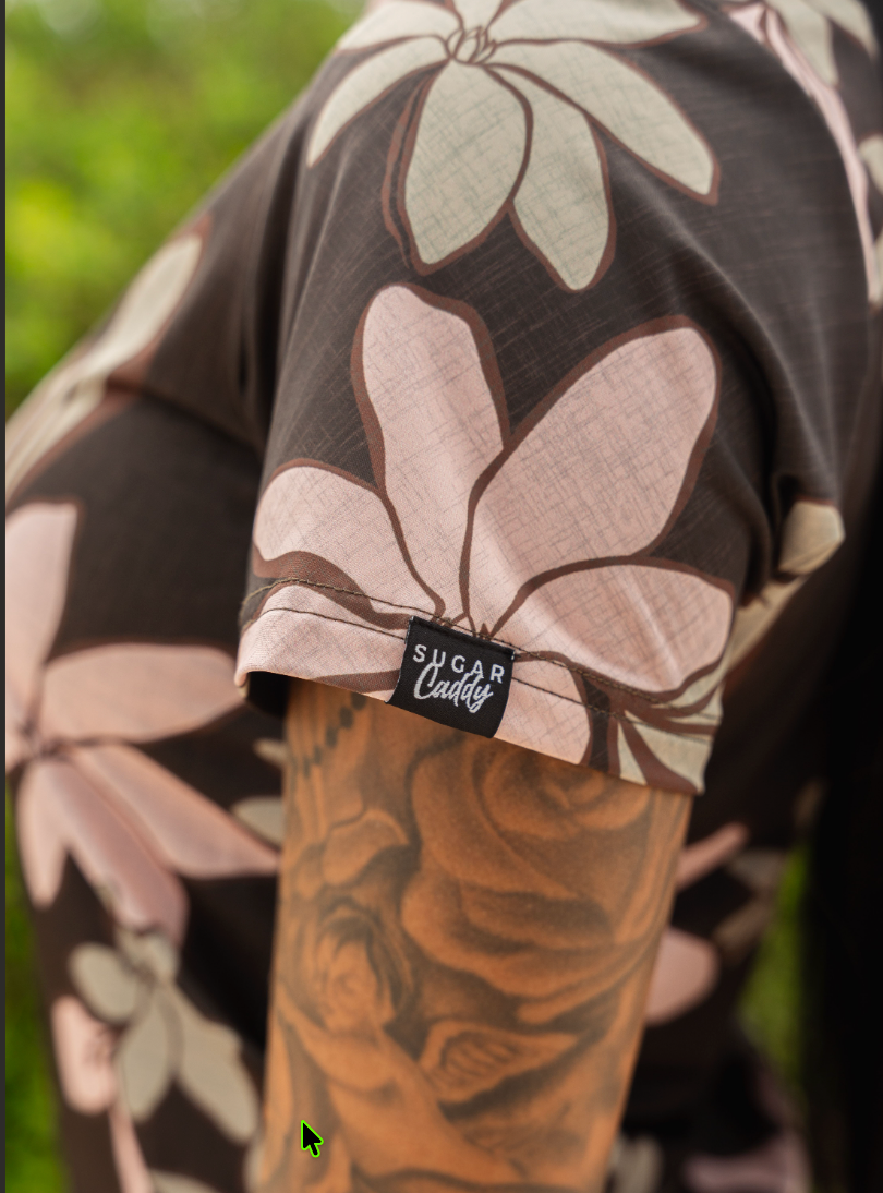 Close-up of a person wearing a dark brown floral shirt with large pale pink and light green flower motifs. A black fabric tag near the cuff reads 'Sugar Cuddie'.