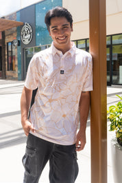 A smiling young man wearing a white short-sleeve polo shirt with a gold abstract floral pattern, standing outdoors in front of a modern building.
