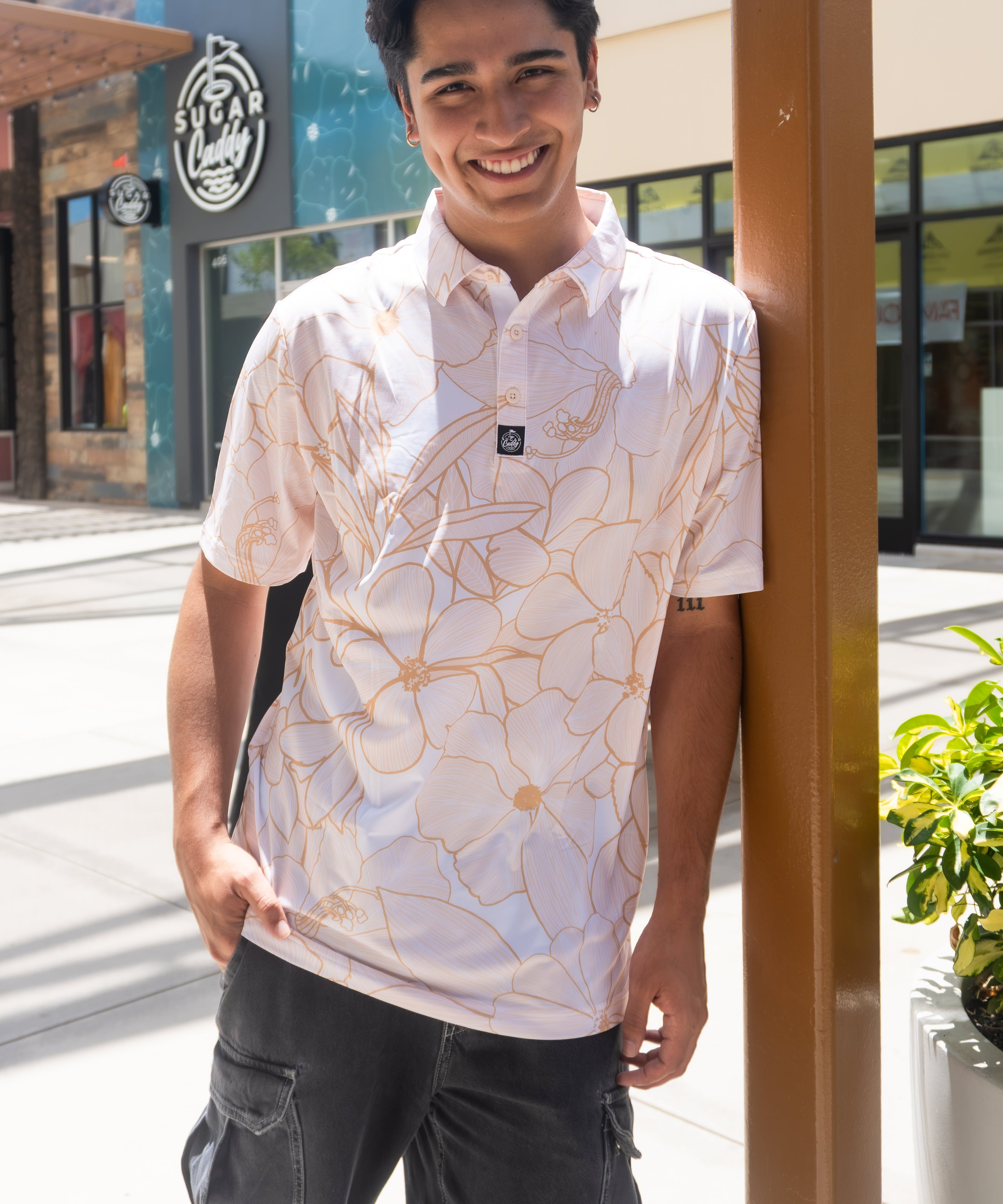 A smiling young man wearing a white short-sleeve polo shirt with a gold abstract floral pattern, standing outdoors in front of a modern building.