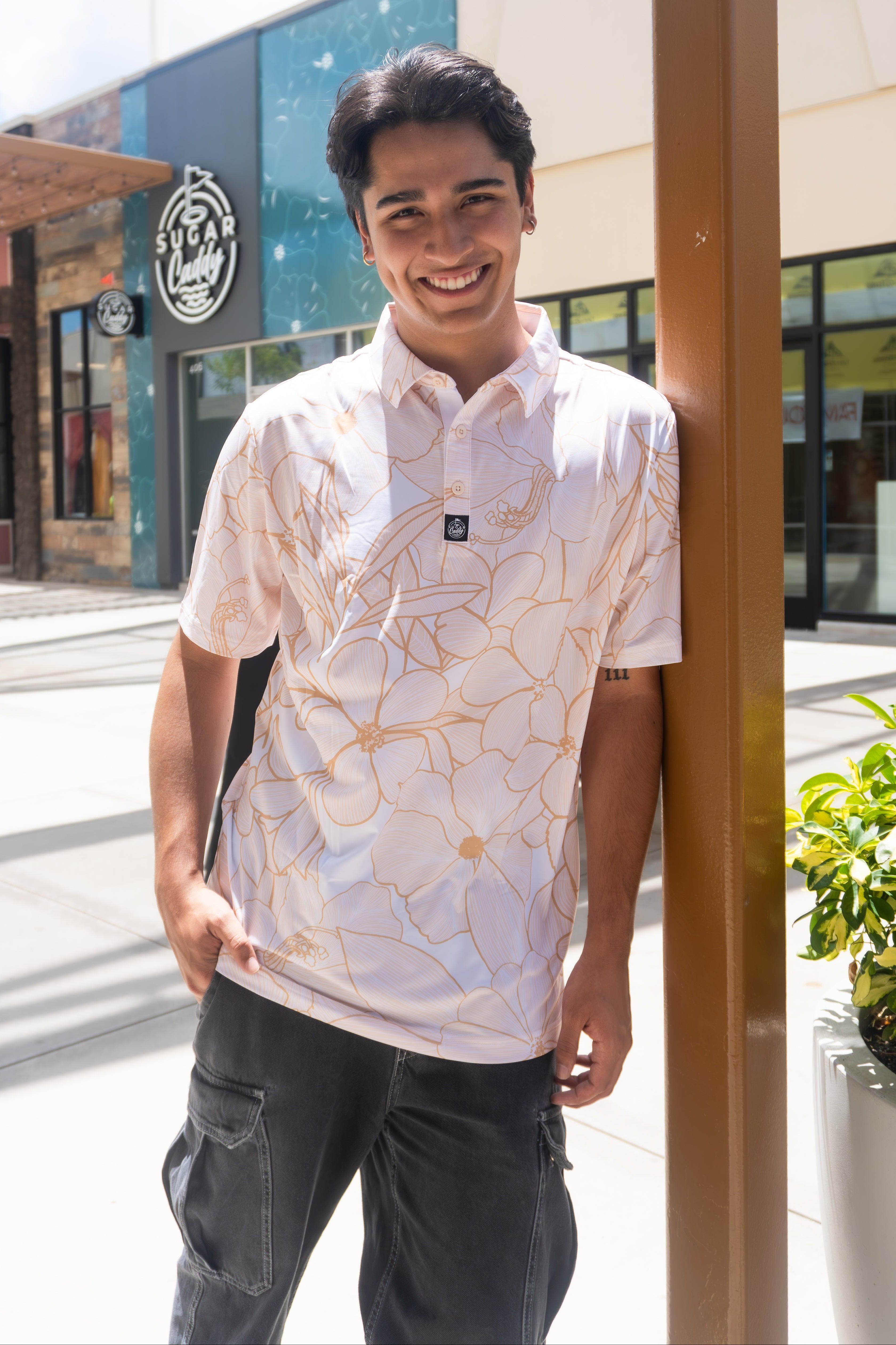 A smiling young man wearing a white short-sleeve polo shirt with a gold abstract floral pattern, standing outdoors in front of a modern building.