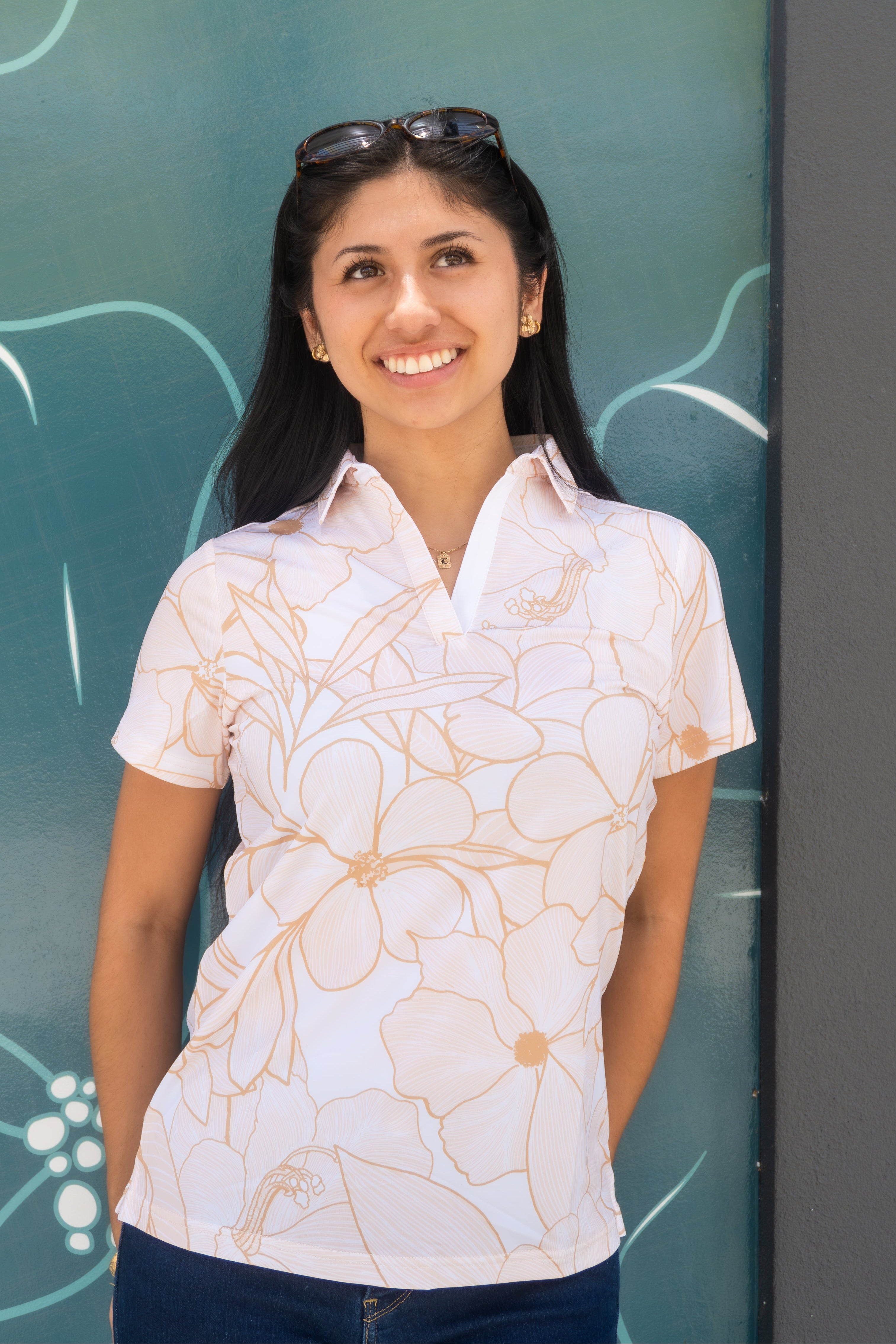 A woman smiling and wearing a white short-sleeve polo shirt with a tan floral pattern, standing in front of a teal decorative wall.
