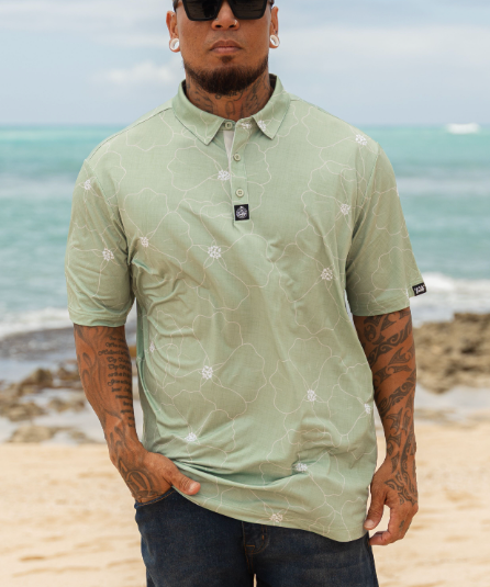 A man wearing a light sage green short-sleeve polo shirt with a white floral pattern, standing on a sandy beach with the ocean in the background.