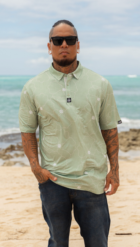 A man wearing a light sage green short-sleeve polo shirt with a white floral pattern, standing on a sandy beach with the ocean in the background.