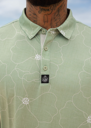 A close-up of a man wearing a light sage green polo shirt with a white floral line pattern and a black embroidered chest logo.