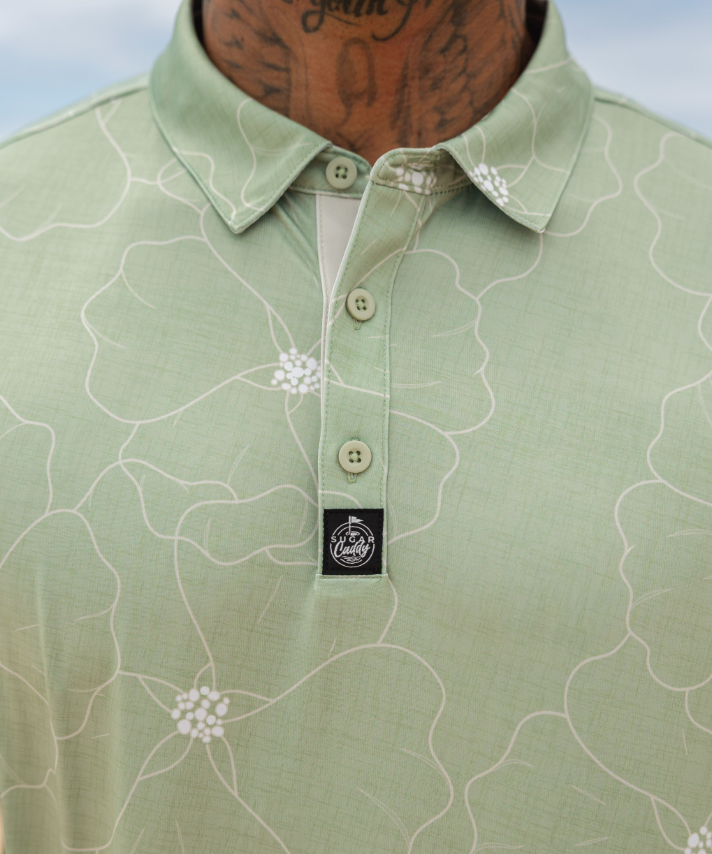 A close-up of a man wearing a light sage green polo shirt with a white floral line pattern and a black embroidered chest logo.