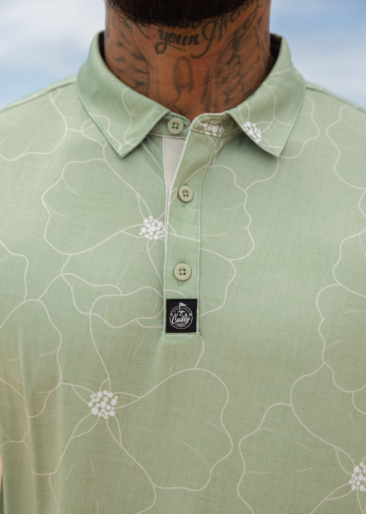 A close-up of a man wearing a light sage green polo shirt with a white floral line pattern and a black embroidered chest logo.