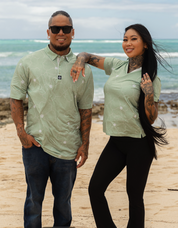 A man and a woman standing on a sandy beach wearing matching light sage green short-sleeve polo shirts with a subtle white floral pattern. The man wears dark blue jeans and sunglasses, and the woman wears black pants.