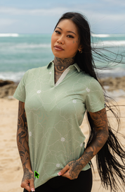 A woman with tattoos stands on a sandy beach wearing a light sage green short-sleeve polo shirt with a white floral pattern. The ocean and sky are blurred in the background.