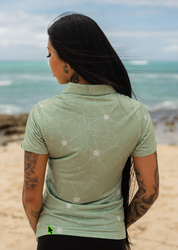 A woman with long black hair stands on a sandy beach with the ocean in the background, wearing a light sage green short-sleeve polo shirt with a white floral pattern on the back.