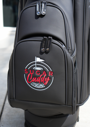A black golf bag with a front pocket featuring a circular 'Sugar Caddy' logo in red and white, photographed outdoors on a light surface.