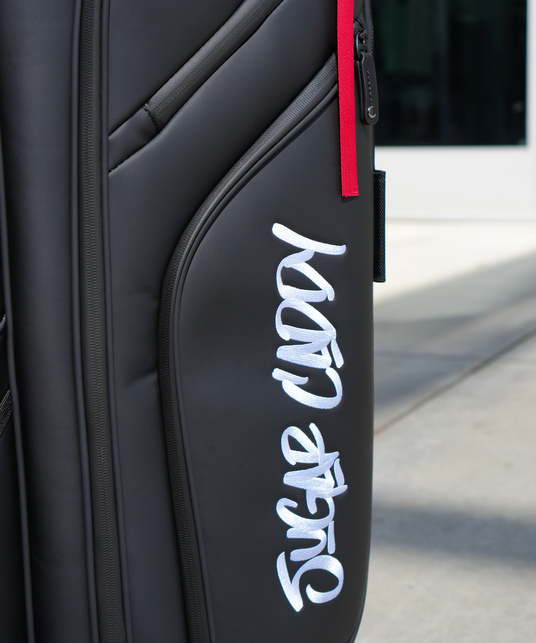 Close-up of a black hard-shell suitcase or duffel bag with a red zipper pull and a large white 'Sugar Caddy' script logo on the front pocket, photographed outdoors on a light concrete surface.