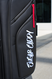 Close-up of a black hard-shell suitcase or duffel bag with a red zipper pull and a large white 'Sugar Caddy' script logo on the front pocket, photographed outdoors on a light concrete surface.